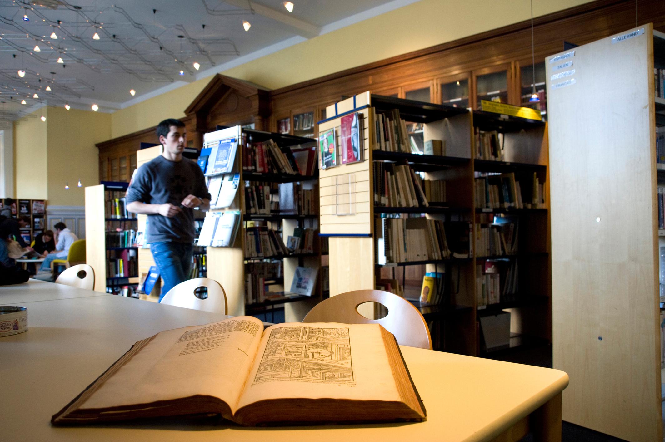 Étudiant parcourant les rayons de la bibliothèque de Mines Saint-Étienne, avec un livre ancien ouvert au premier plan.