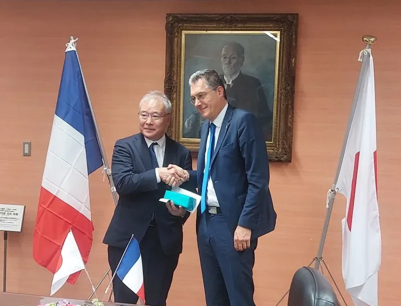 Poignée de main entre le directeur de Mines Saint-Étienne et un représentant japonais lors de la signature d'un partenariat éducatif, avec les drapeaux français et japonais en arrière-plan.